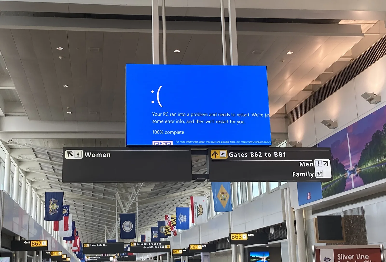 An airport screen showing the Windows blue error screen above signs for gates B62 to B81