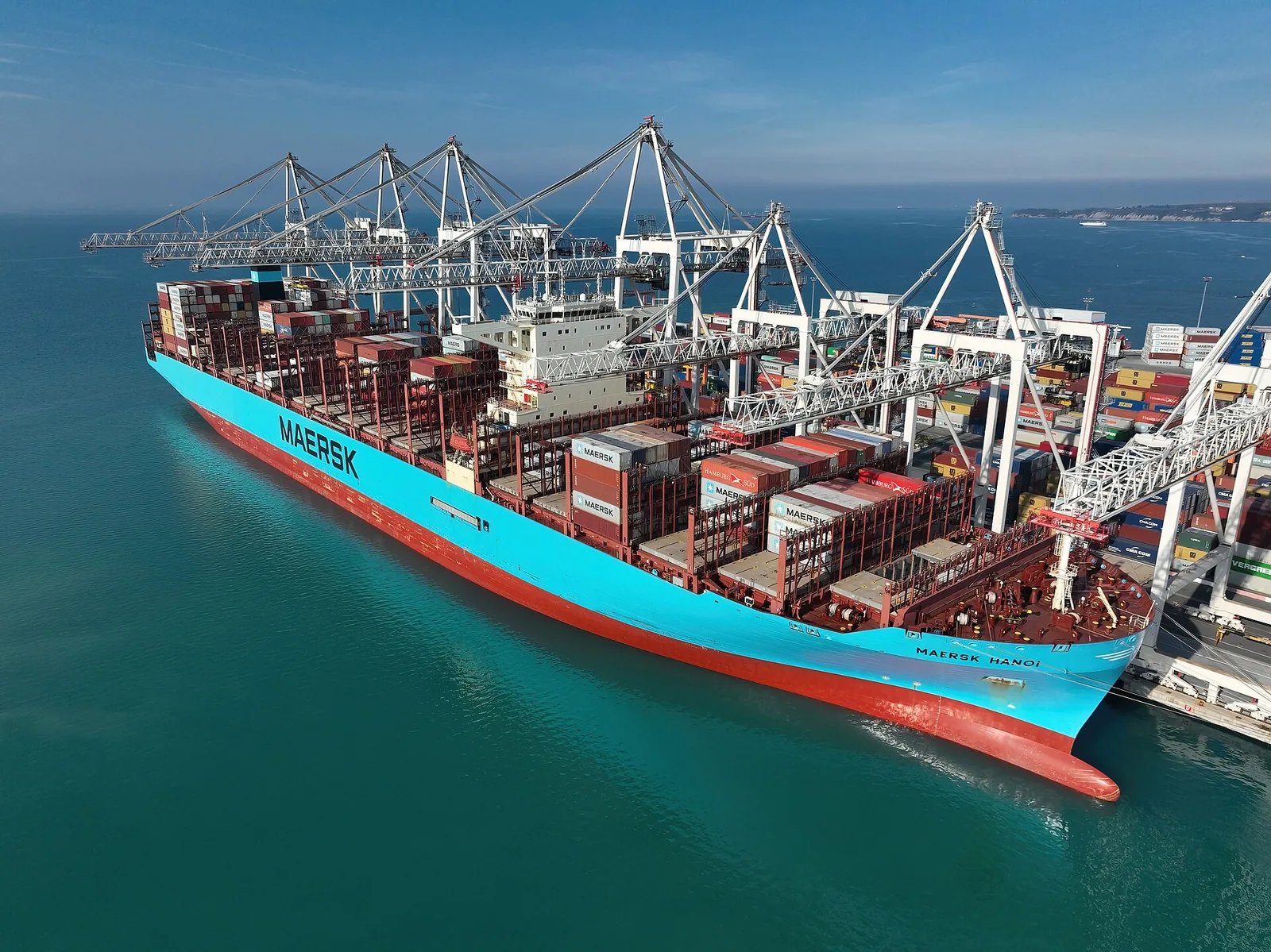 A large Maersk container ship loaded with containers, berthed under port cranes, seen from above