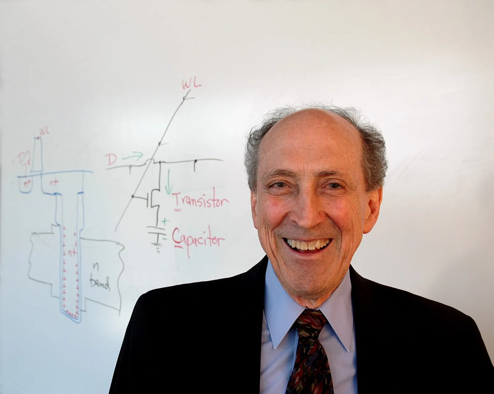 Robert Dennard smiling in front of a whiteboard sketch of a transistor and a capacitor