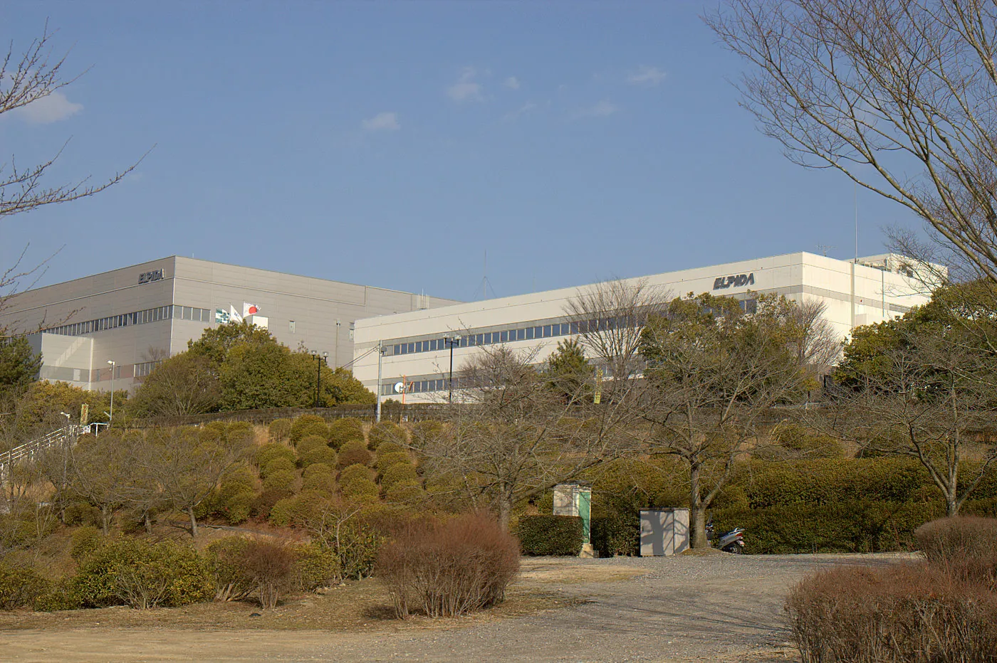 Low white factory buildings with an Elpida sign behind a garden
